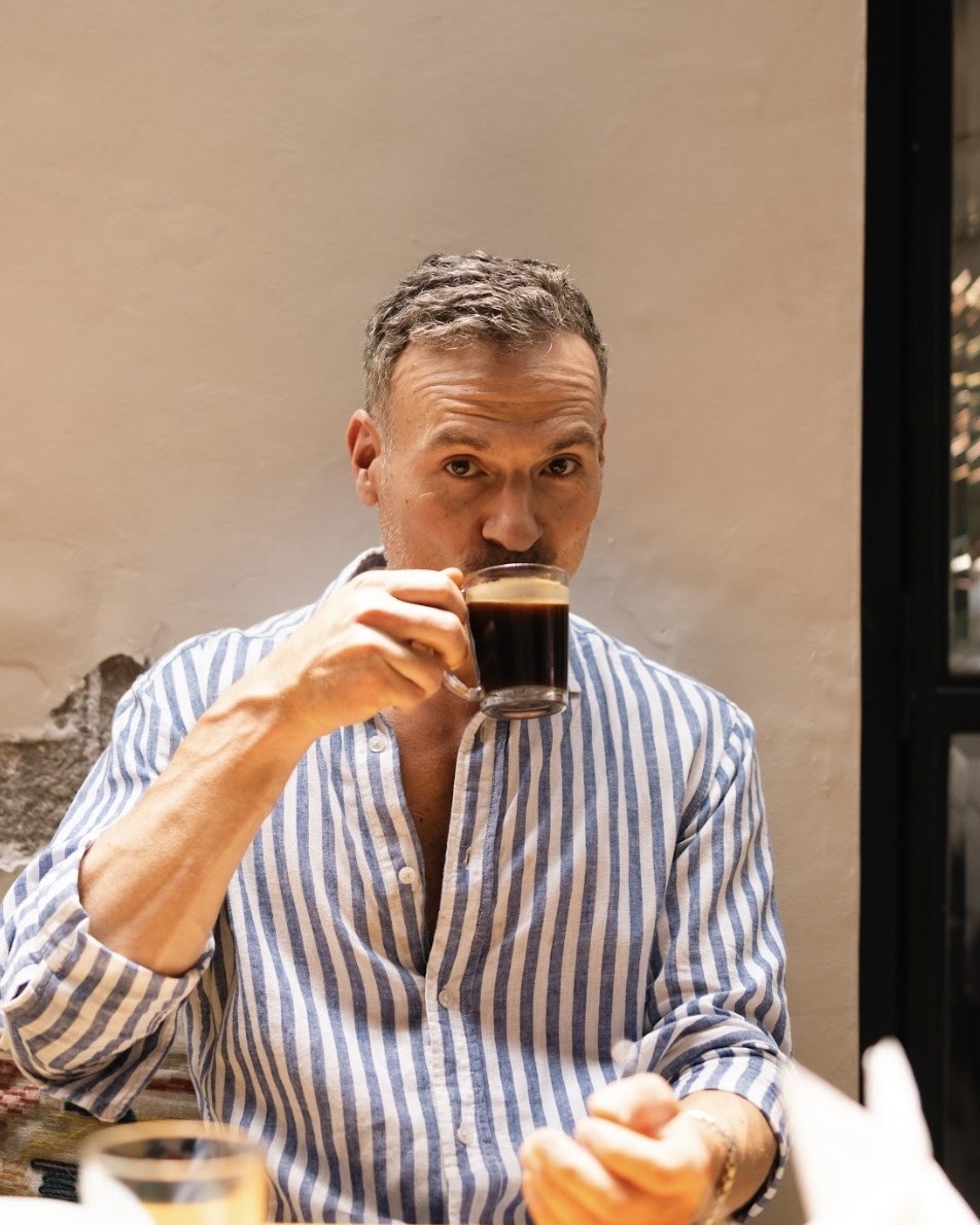 Ray, photographer, seated at a caf&eacute; table drinking an espresso against a pale plaster wall